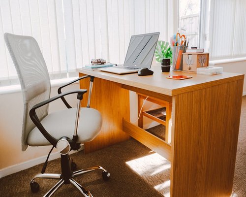 A well-organized wooden home office desk with an ergonomic black mesh office chair facing a brightly lit window